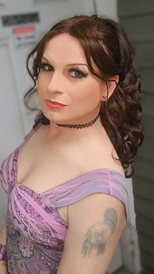 A picture of Heather Thomas. A stunningly beautiful woman of the caucasian persuasion
         with dark hair pulled up into a ponytail. She is wearing purple heart stud earrings, a black choker, a purple paisley summer dress,
          and makeup to slay the gods. There is a tattoo of a skull with a ponytail and a eastern style moustache and beard that she regrets
          and will someday get covered up. The background is a door with some kind of warning sign on it and the side of a house. The background
           is blurred out, so the focus is on her.